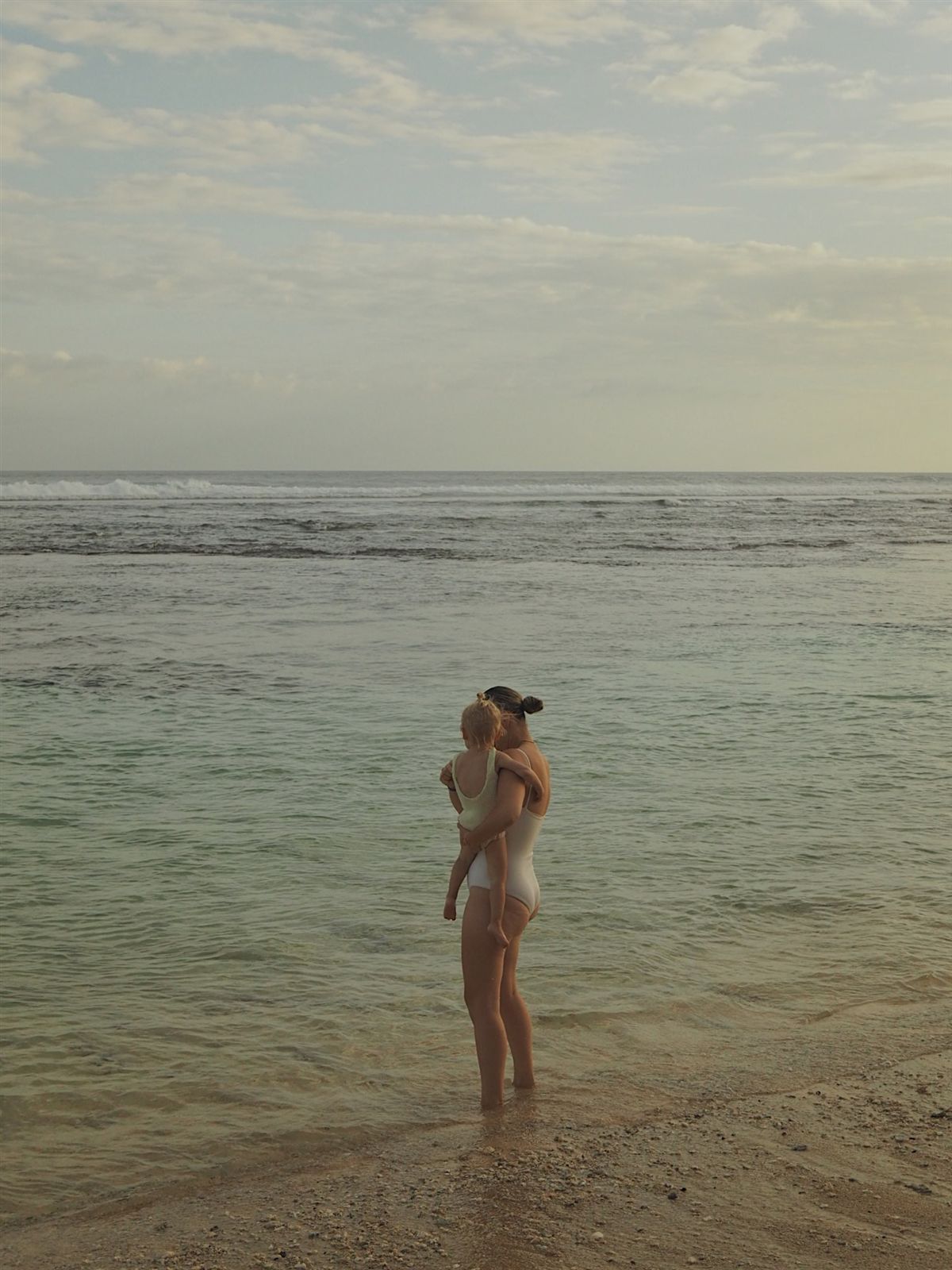 A mother holding her toddler while standing in shallow ocean water at sunset